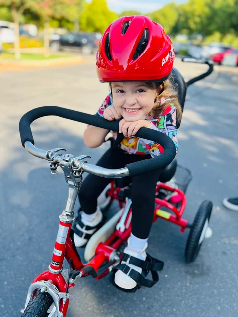 A young girl in a red helmet grins over the handlebars of her red adaptive trike.