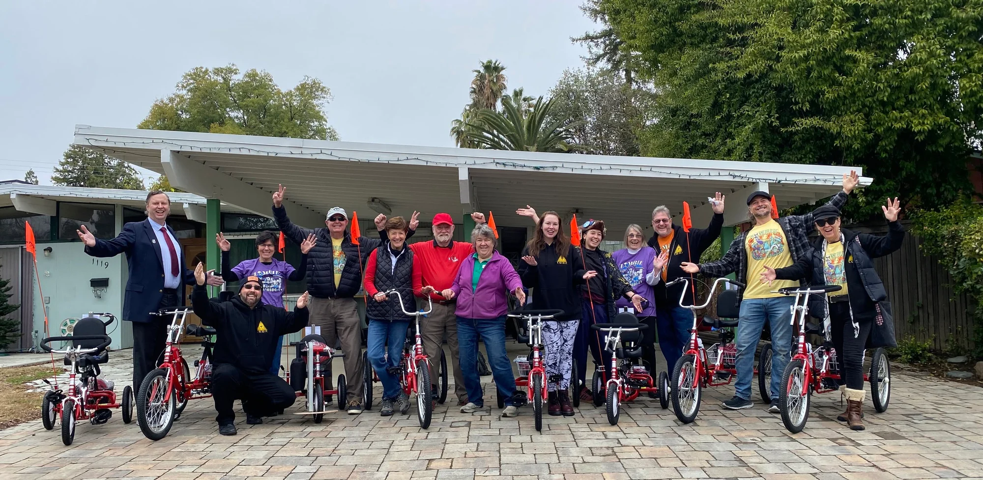 A group stands cheering with arms raised behind a row of adaptive trikes outside a workshop.