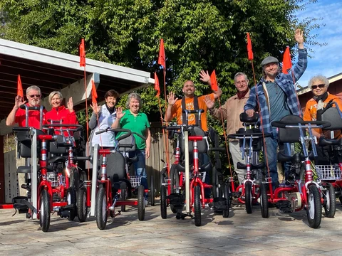 A line of people wave from behind a row of red adaptive trikes with orange safety flags.