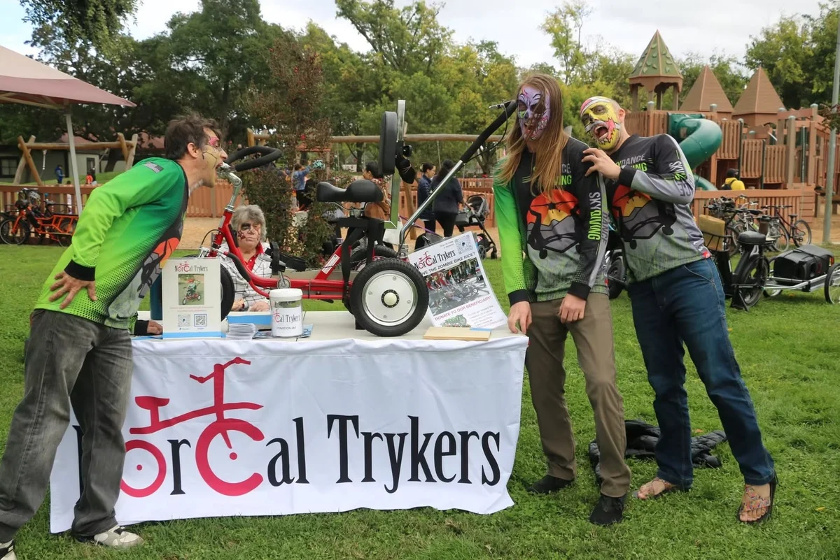 Costumed festival-goers lean over the NorCal Trykers information table on the park lawn.