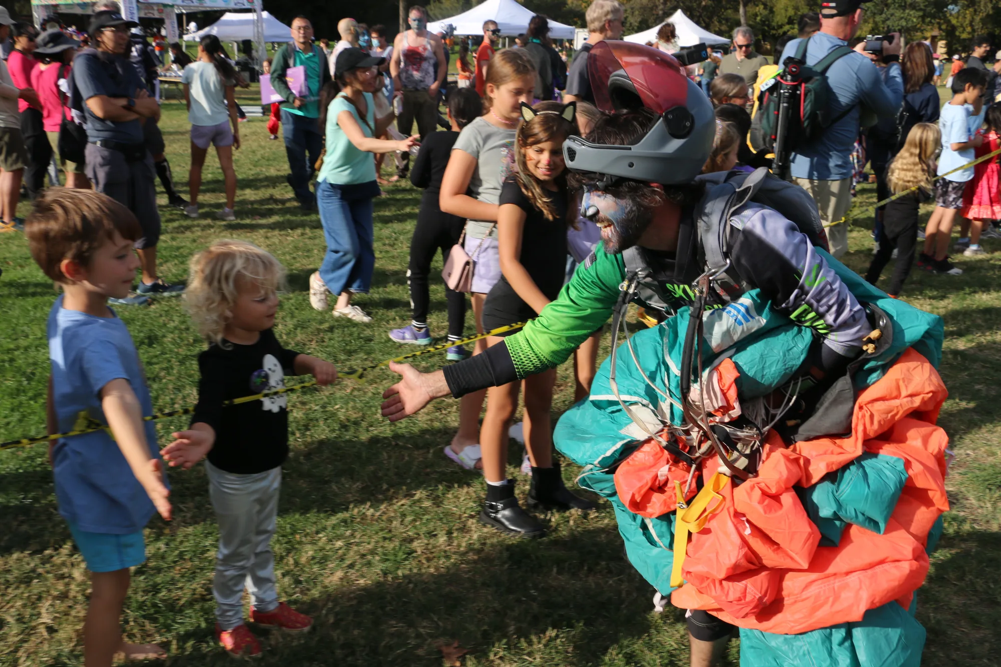 A costumed skydiver in a red and teal parachute rig greets a young child.